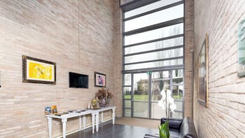 Un hall d’entrée spacieux avec un plafond haut, des murs en briques et de grandes fenêtres. On y trouve un banc blanc, un canapé en cuir noir et une petite table sur laquelle est posée une plante en pot.