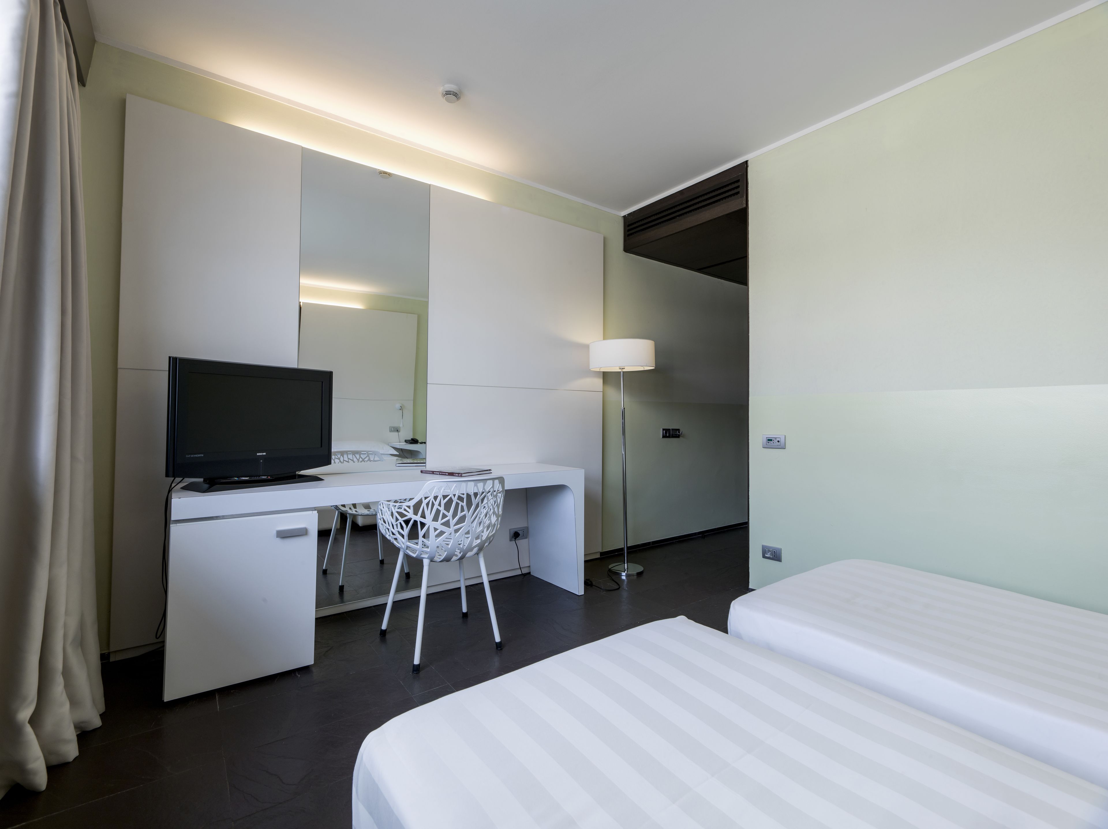A hotel room with a bed, a desk with a computer, a chair, a lamp, and a television.