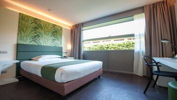 Et hotelværelse med en seng, et skrivebord, en stol, et vindue med gardiner og en væg med bladmotiv.