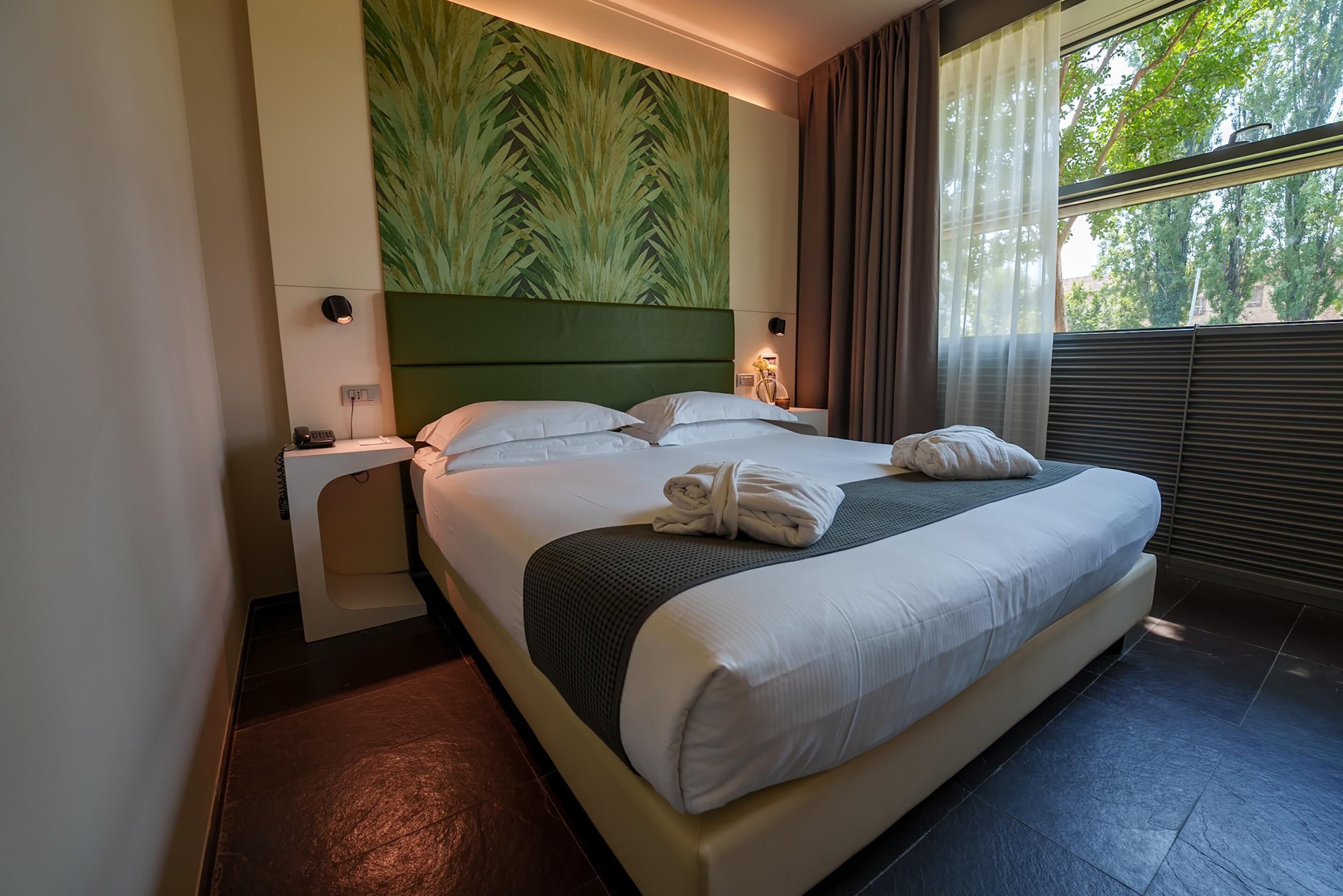 A hotel room with a large bed, a headboard with a leaf pattern, bedside lamps, and a window with curtains.