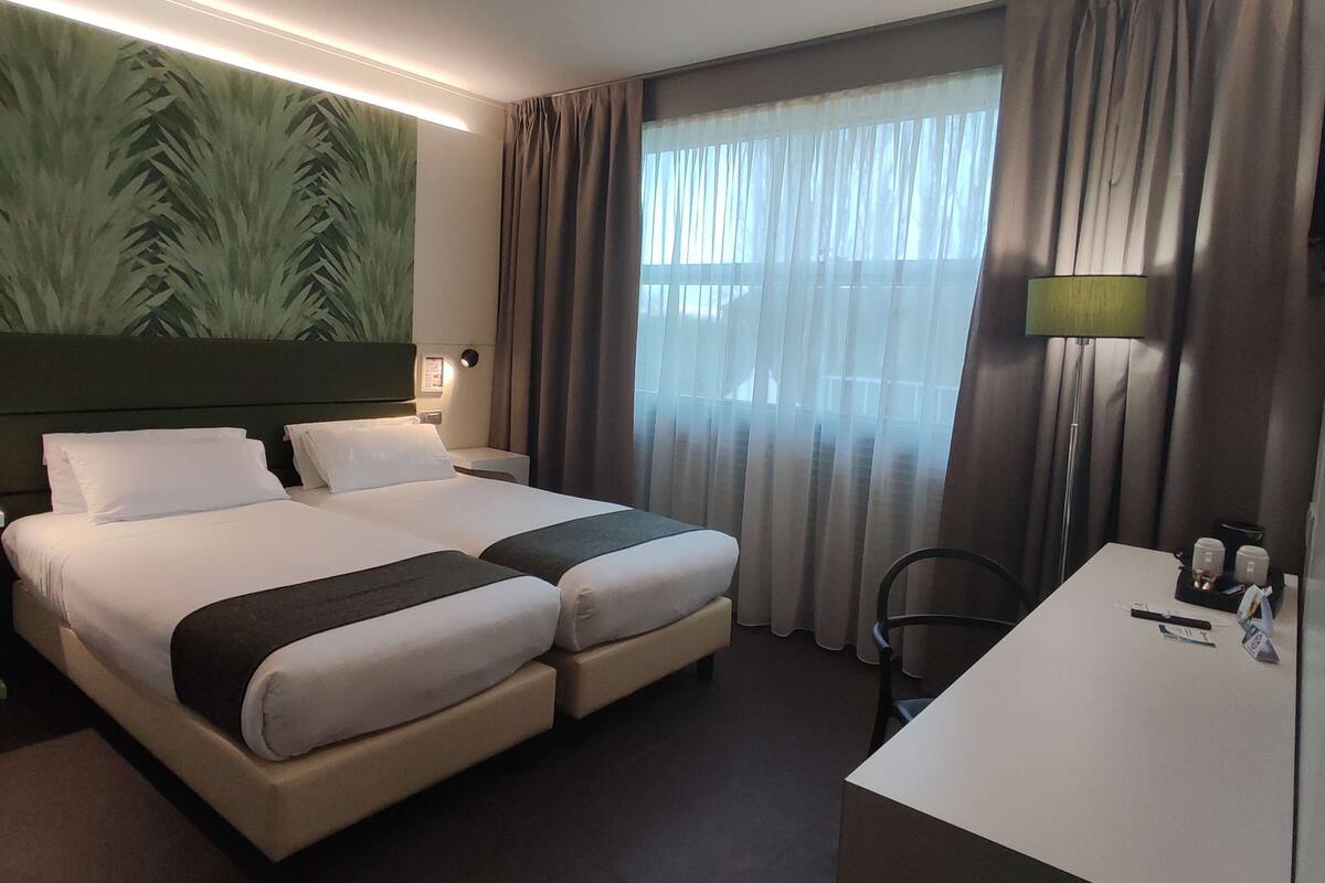 A hotel room with two beds, a desk, a lamp, and a large window with curtains.