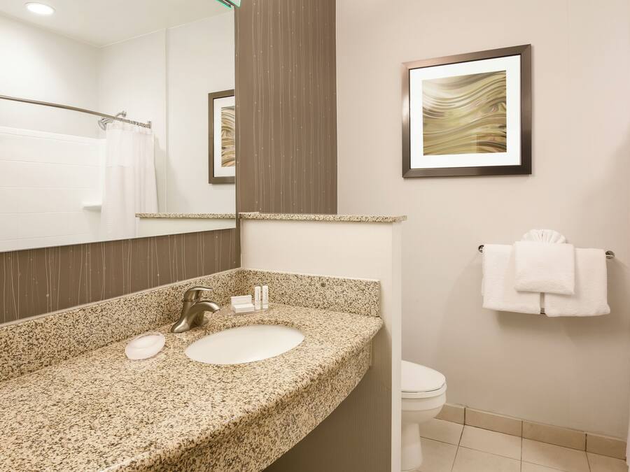 Une salle de bain avec un lavabo doté d’un plan de travail en granit, un miroir, une œuvre d’art encadrée, un porte-serviettes et des toilettes.