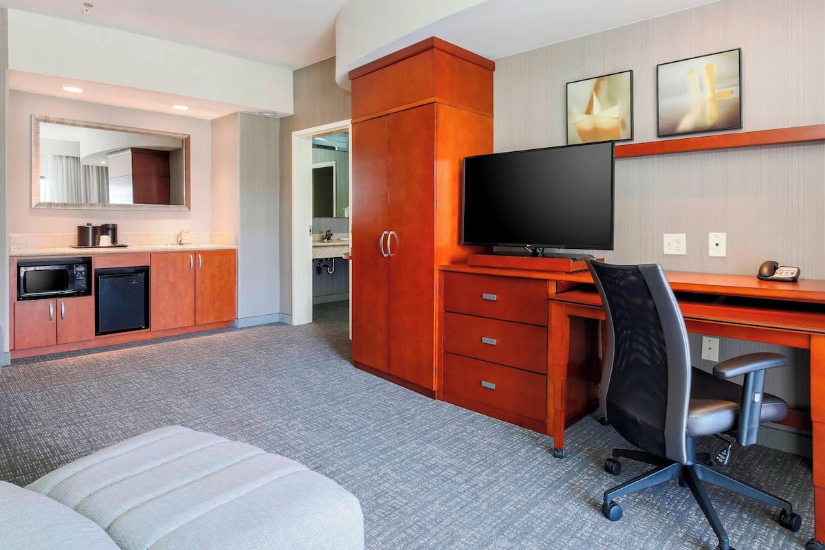 A hotel room with a kitchenette, a large TV, a desk with a chair, and a large mirror.