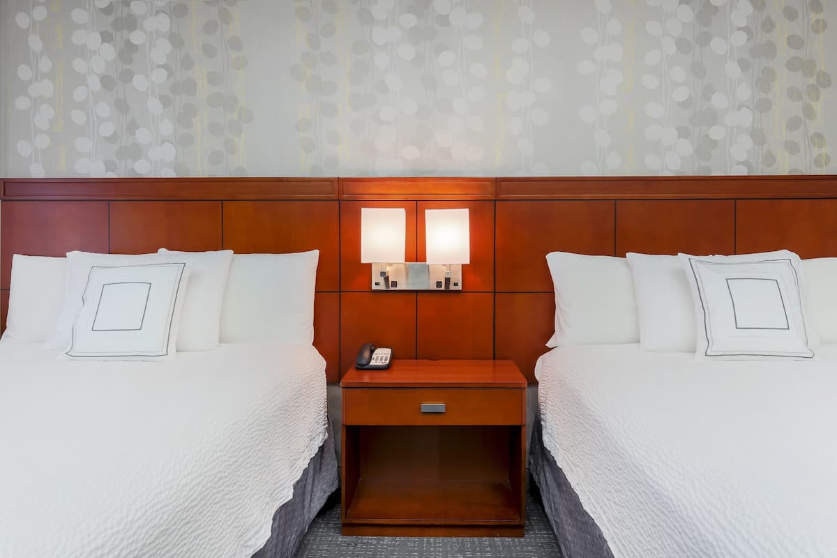 A hotel room with two beds, a wooden headboard, a nightstand with a phone, and wall-mounted lights.