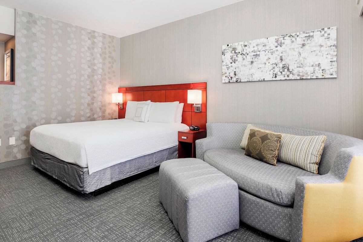 A hotel room with a bed, a gray sofa, a nightstand, and a wall-mounted artwork.
