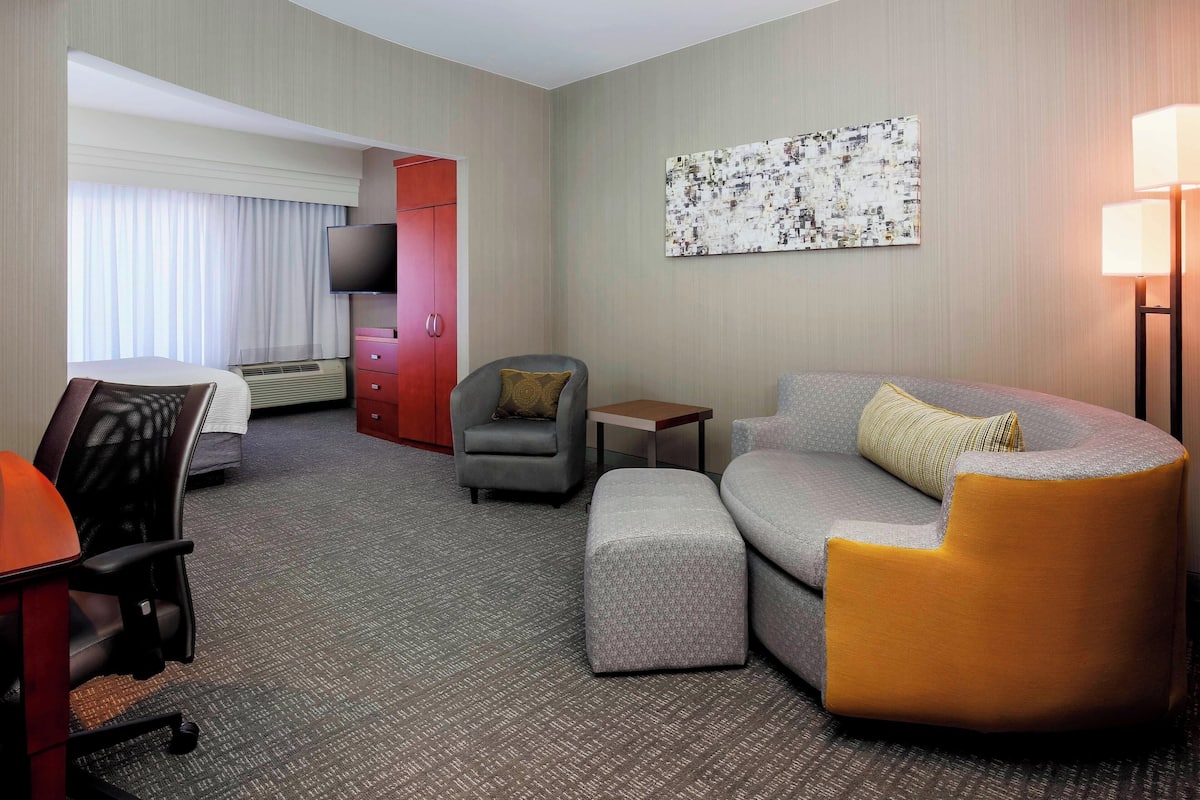 A hotel room with a grey sofa, a small armchair, a desk with a chair, a flat-screen TV, a red wardrobe, and a large abstract painting on the wall.