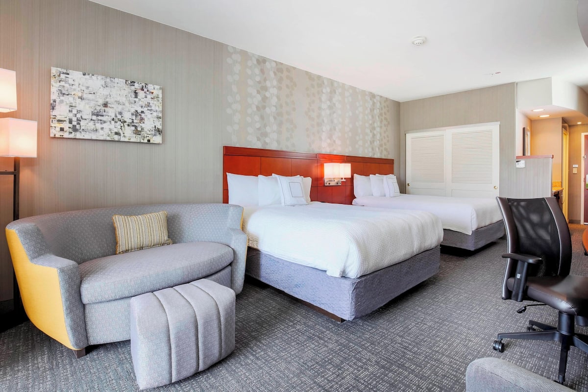 A hotel room with a gray sofa, two single beds, a desk with a chair, and a large window.