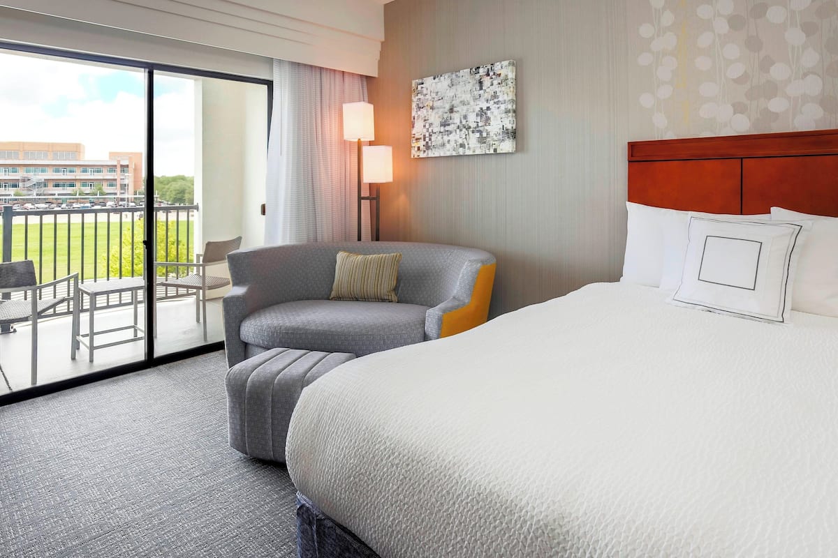 A hotel room with a large bed, a grey armchair, a small table, and a view of the outside.