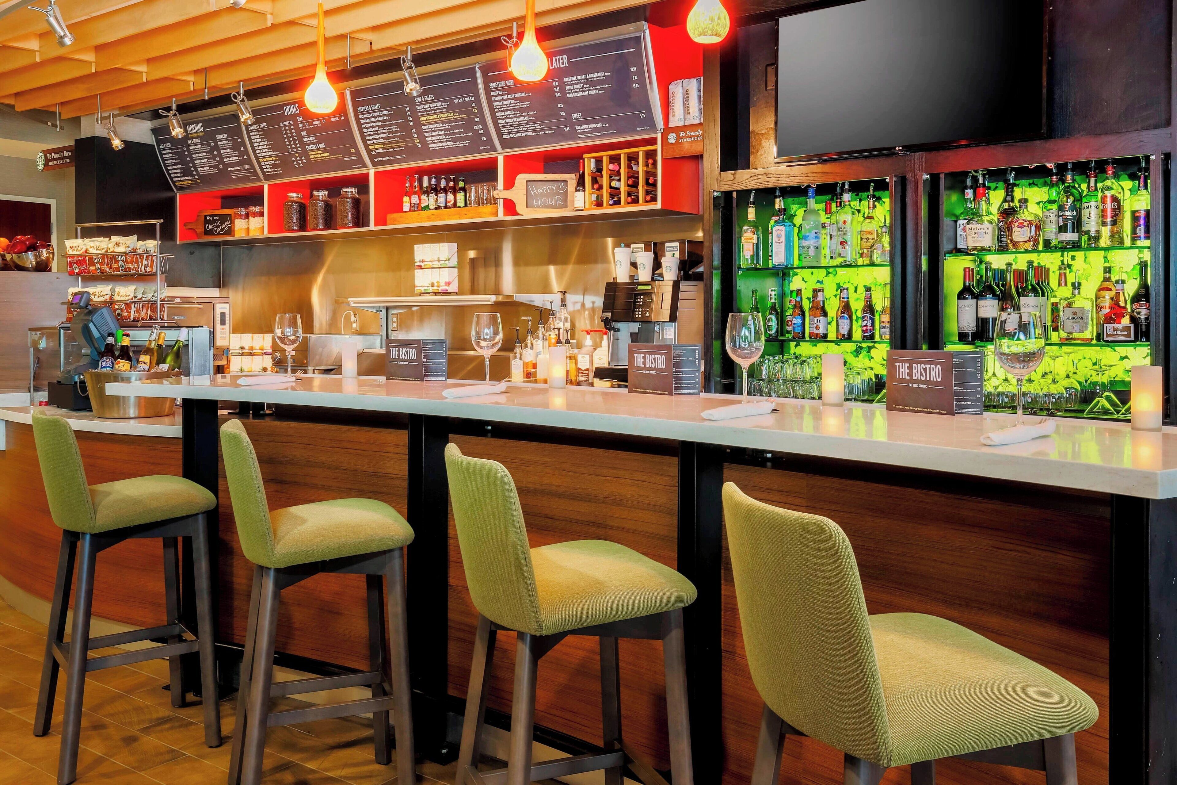 A bar area with a wooden counter, green bar stools, a menu board, and a variety of liquor bottles.