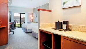 Ein Hotelzimmer mit Bett, Schreibtisch mit Kaffeemaschine, Stuhl und Blick auf ein Gebäude durch ein großes Fenster.