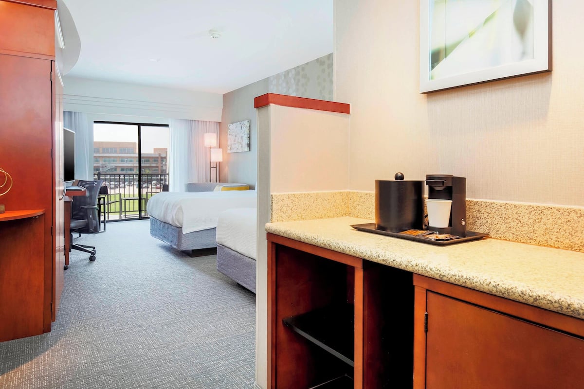 A hotel room with a bed, a desk with a coffee maker, a chair, and a view of a building through a large window.