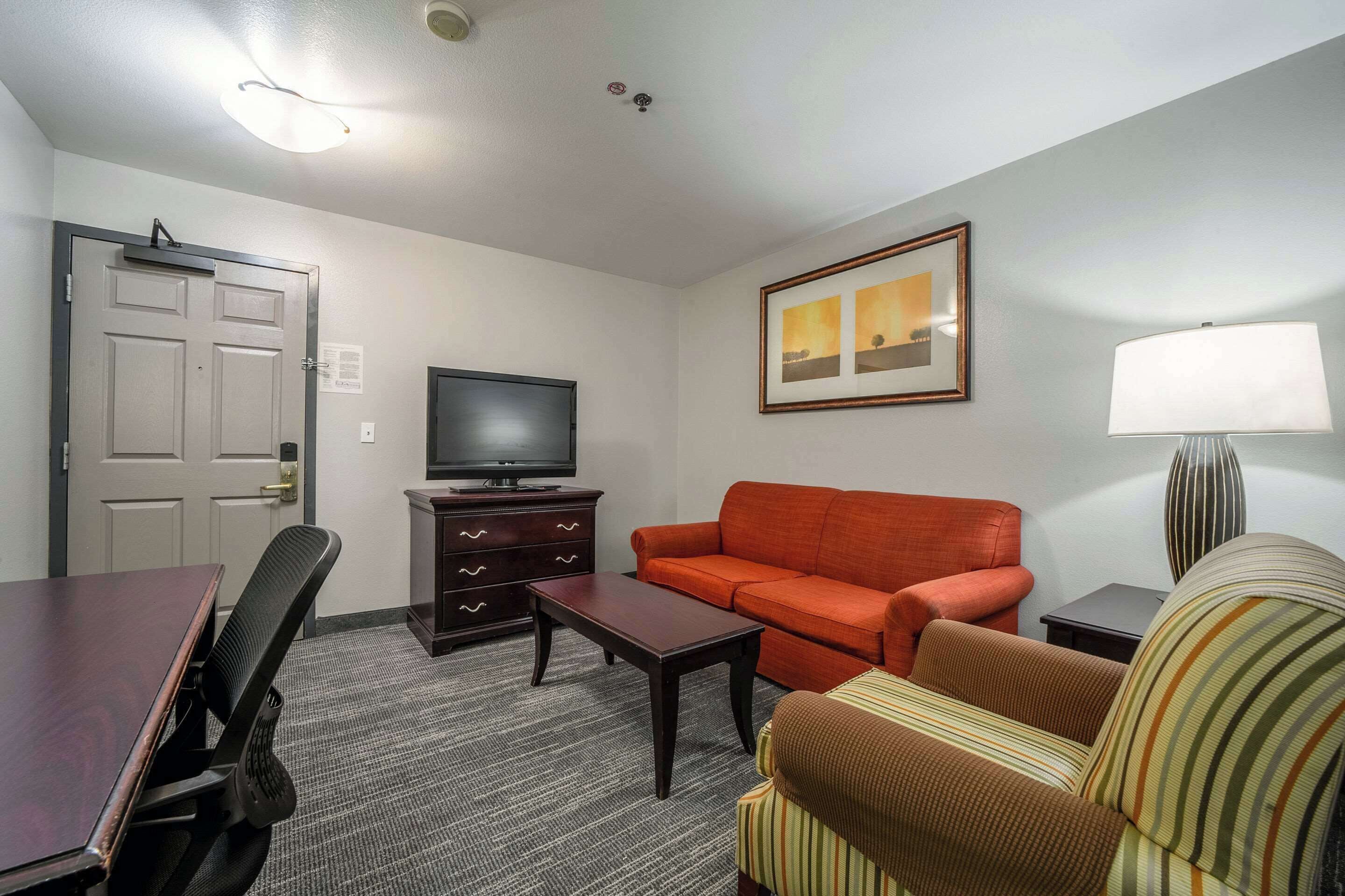 A hotel room with a television, a sofa, a striped armchair, a small table, a lamp, and a door.