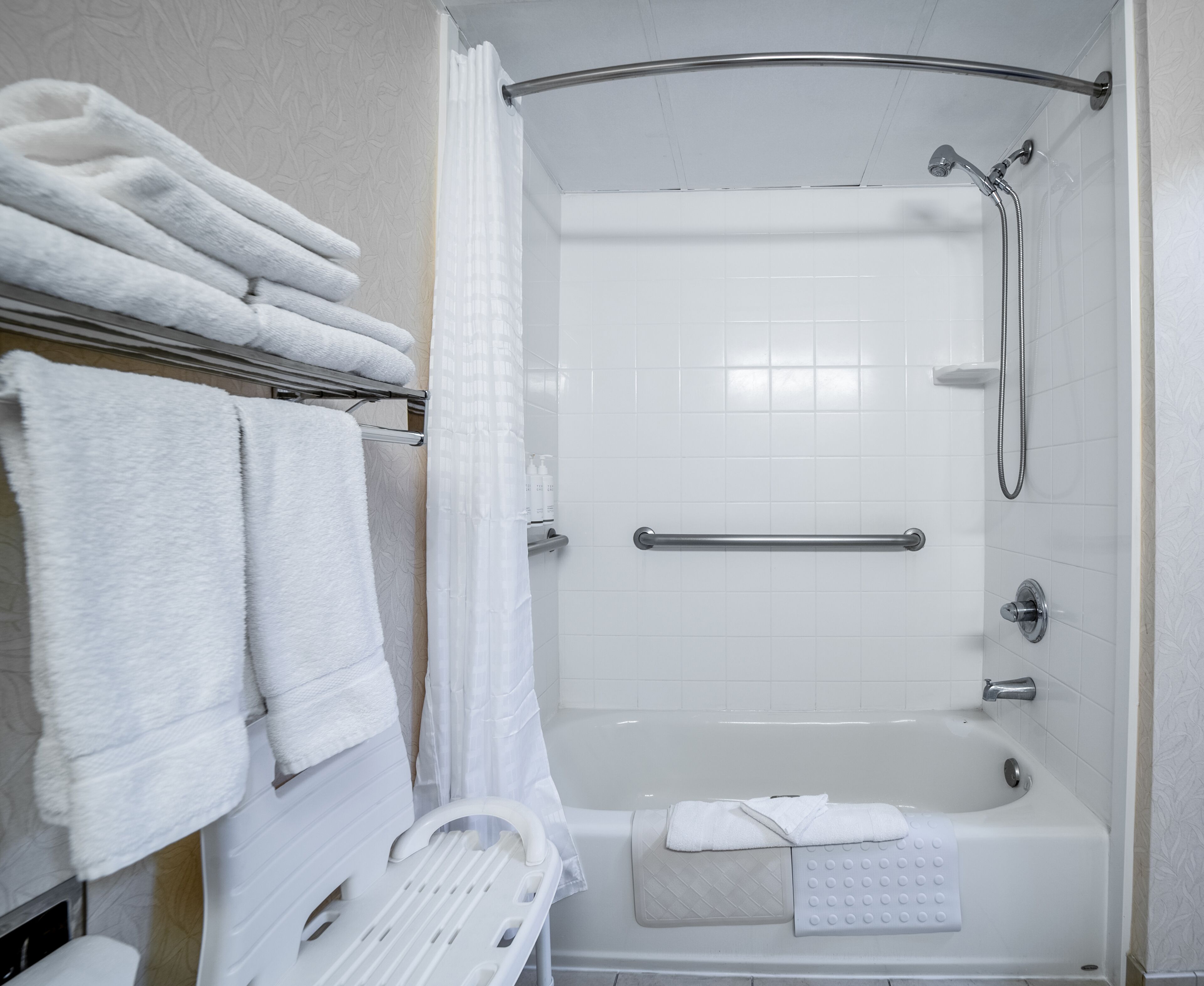 A modern bathroom with a shower, a bathtub, and a handrail. There are multiple white towels on a rack and a shower caddy.