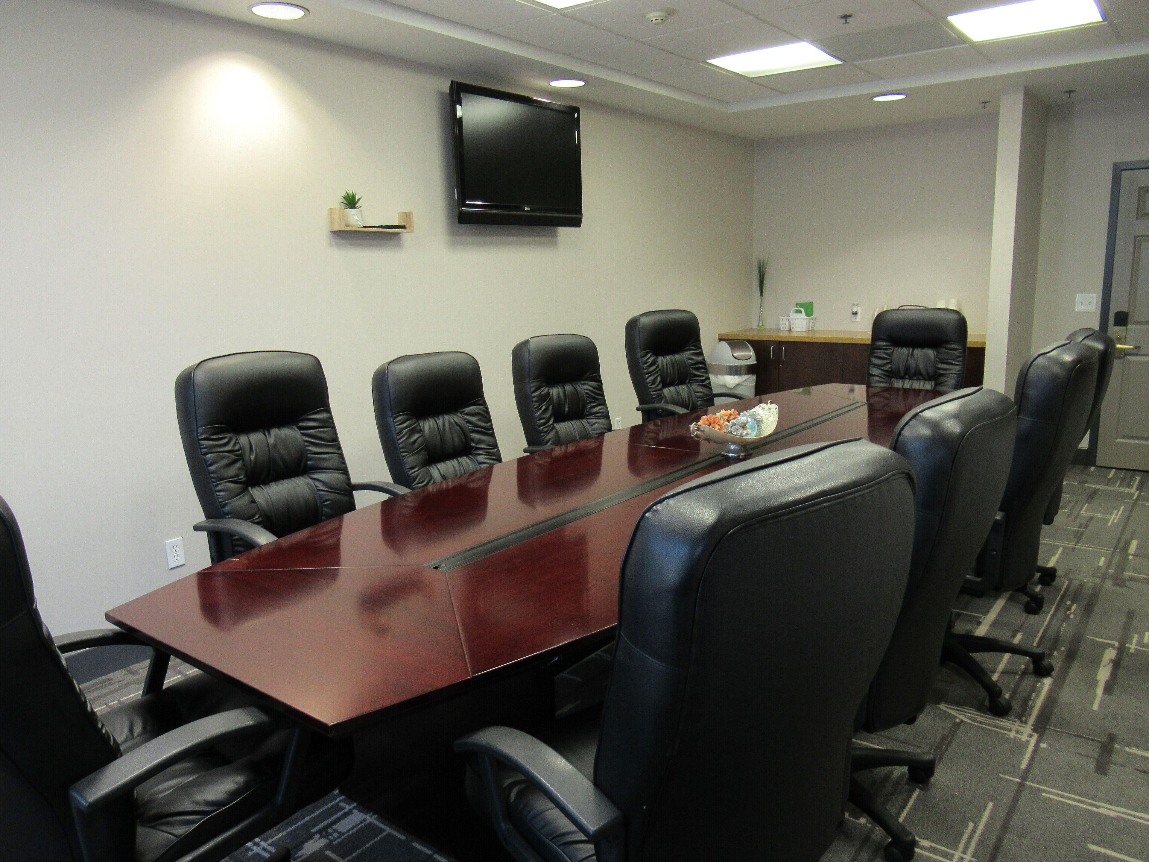 A conference room with a long table, six black leather chairs, a flat-screen TV on the wall, and a small plant on a shelf.