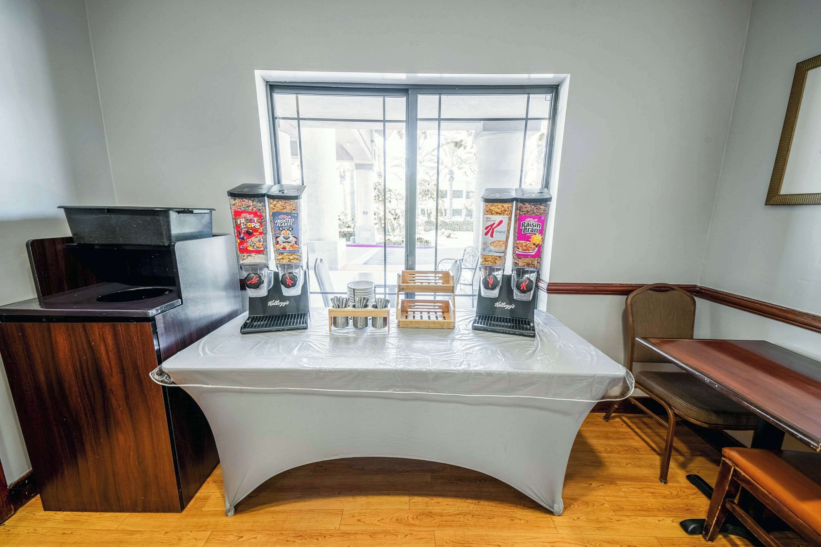 Una habitación con una mesa cubierta por un mantel blanco, dos cafeteras, un banco de madera y una ventana con vista al exterior.