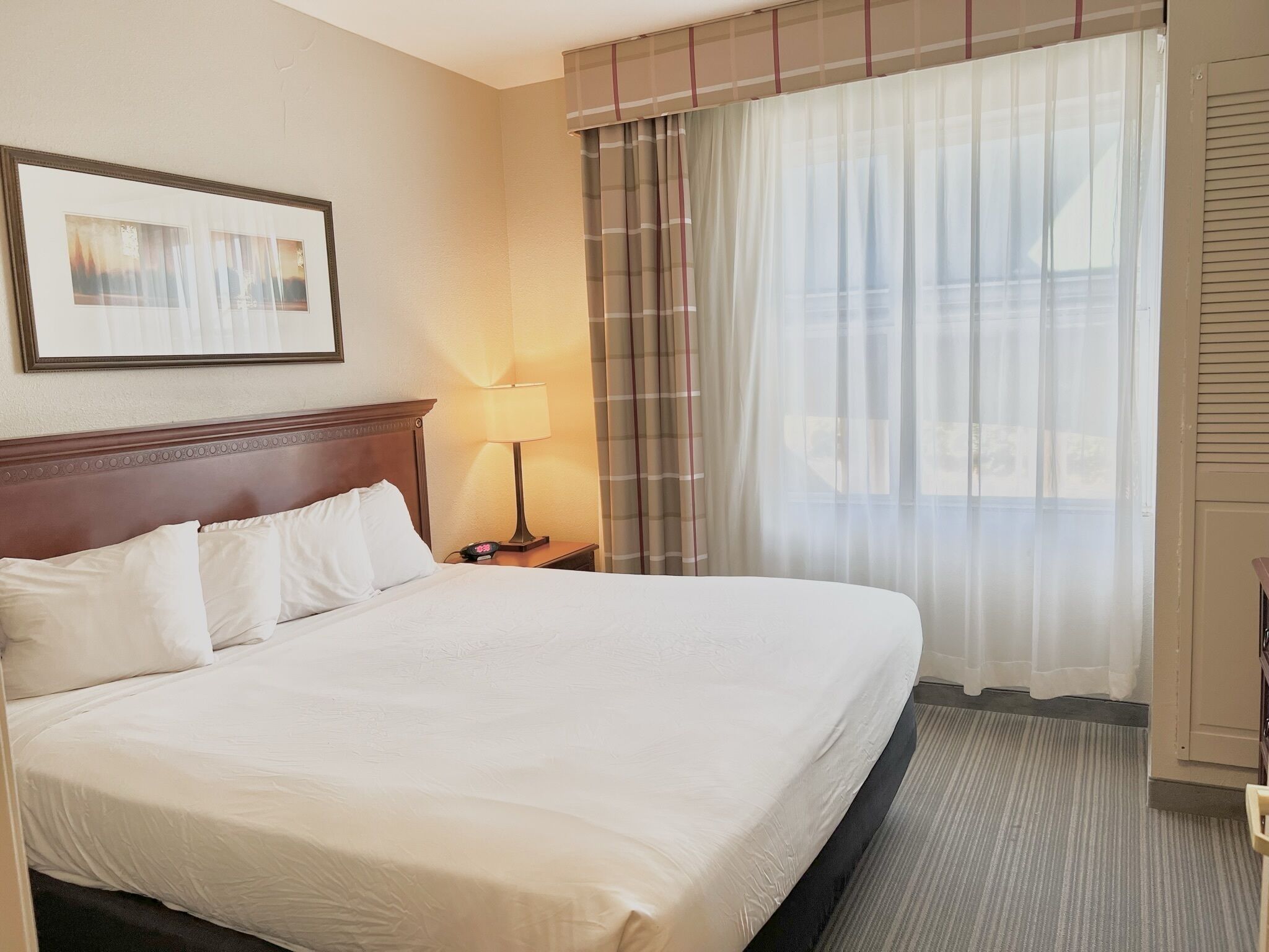 Habitació d'hotel amb un llit gran, una tauleta de nit amb una làmpada, un quadre amb marc a la paret i una finestra amb cortines translúcides.