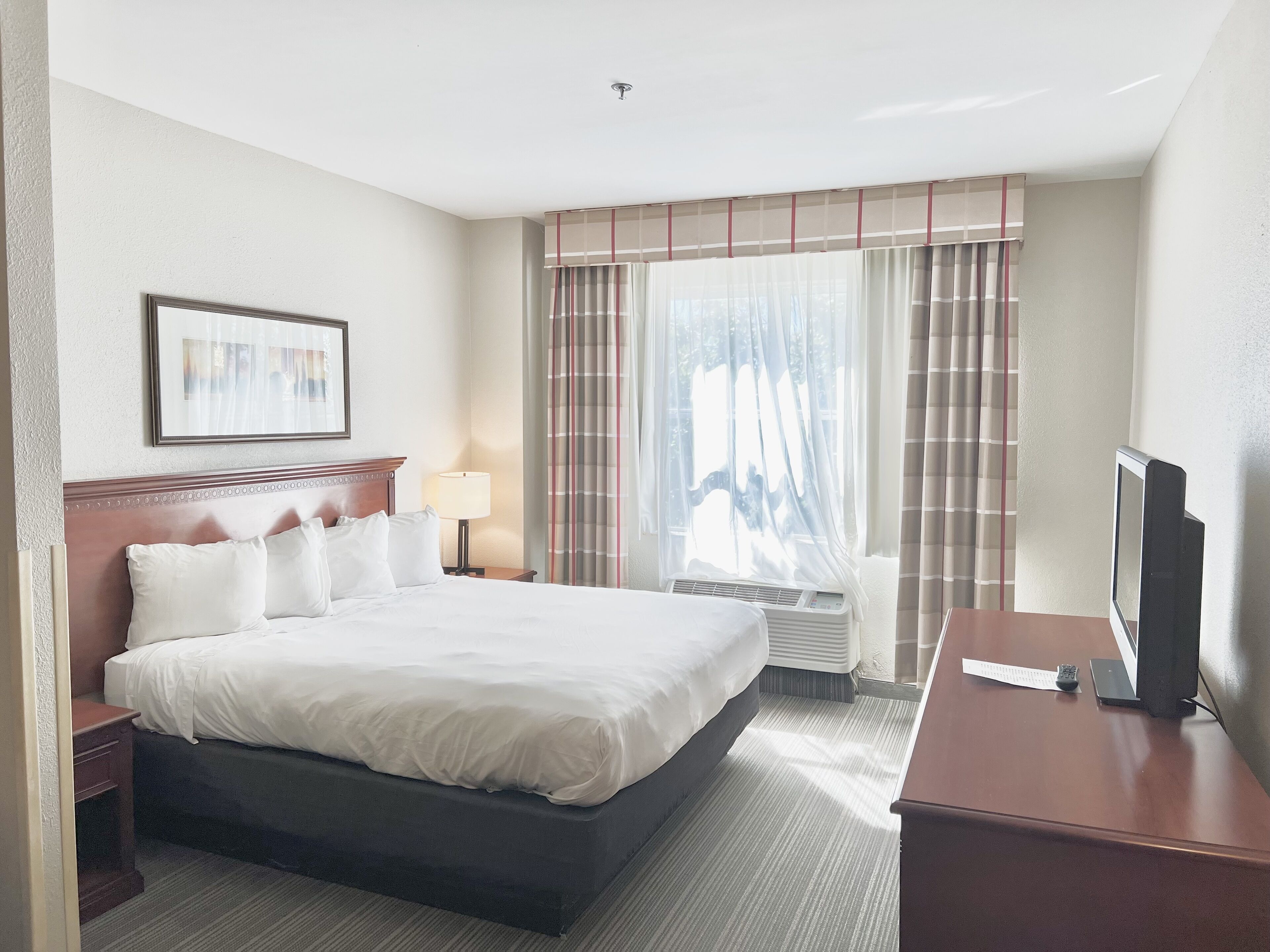 A hotel room with a large bed, a bedside table with a lamp, a TV on a stand, and a window with curtains.