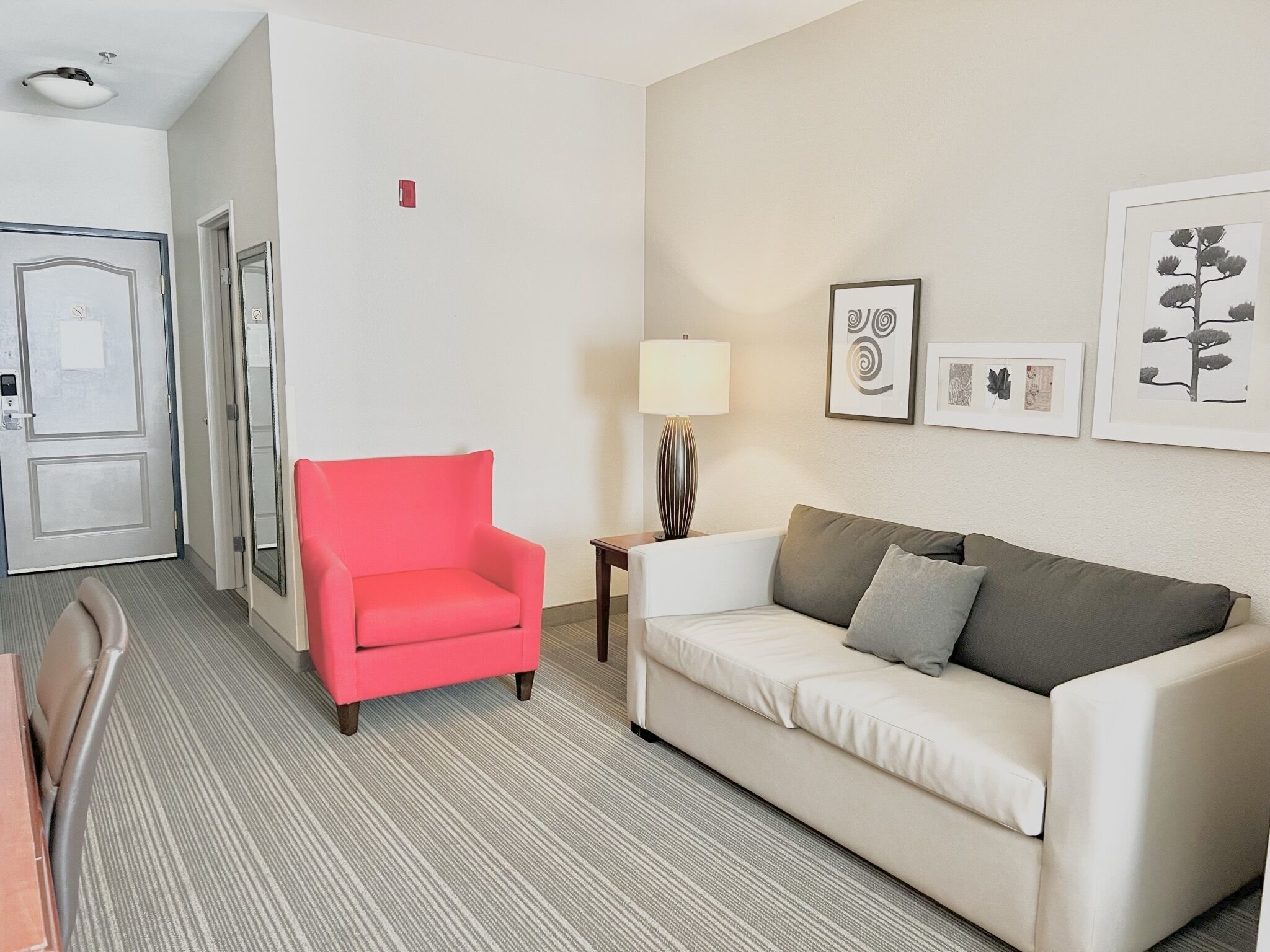 A hotel room with a red armchair, a beige sofa, a small table with a lamp, and framed botanical prints on the wall.
