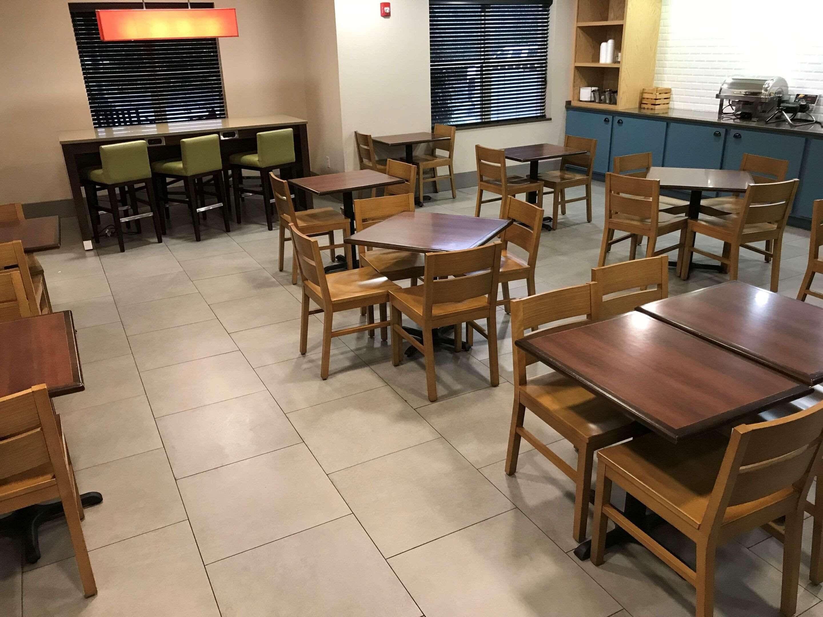 A dining area with multiple wooden tables and chairs, a kitchen area with blue cabinets, and a window with blinds.