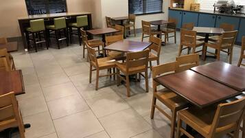 A dining area with multiple wooden tables and chairs, a kitchen area with blue cabinets, and a window with blinds.