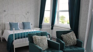 Un dormitorio con cama, dos sillones, una mesita de noche y una ventana con cortinas.
