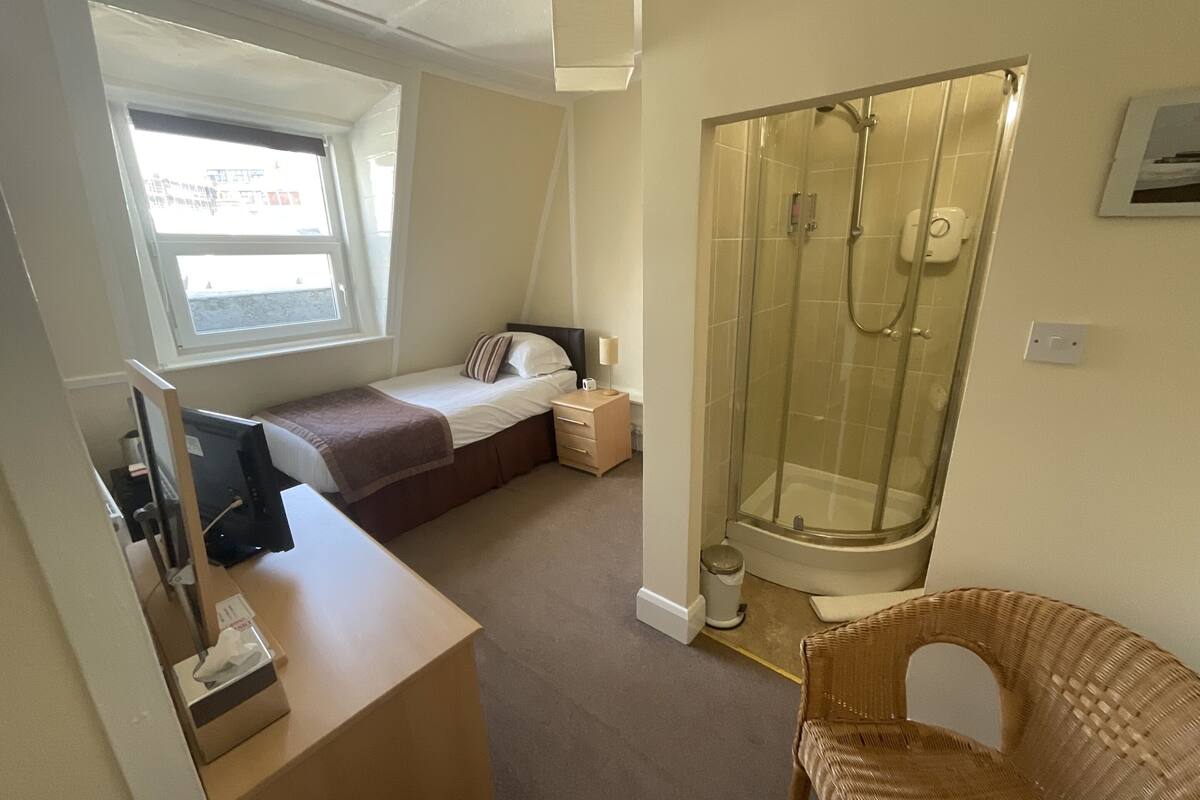 A bedroom with a bed, a bedside table, a window, a shower, and a chair.