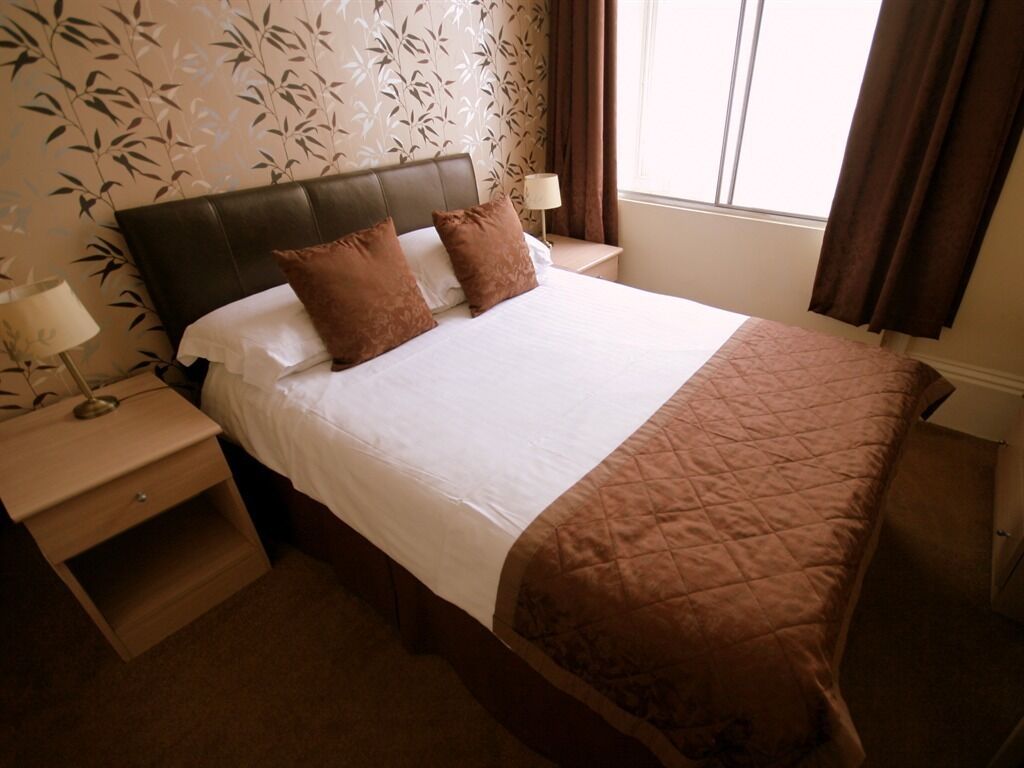 Cama bem arrumada com colcha marrom, dois travesseiros e uma mesa de cabeceira de madeira com abajur.