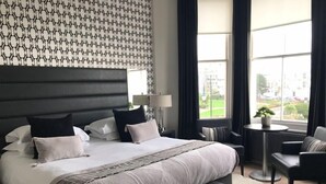 A hotel room with a large bed, a bedside table with a lamp, a small round table with a plant, and a view of the outside through large windows. - Hastings House (St Leonards-On-Sea)