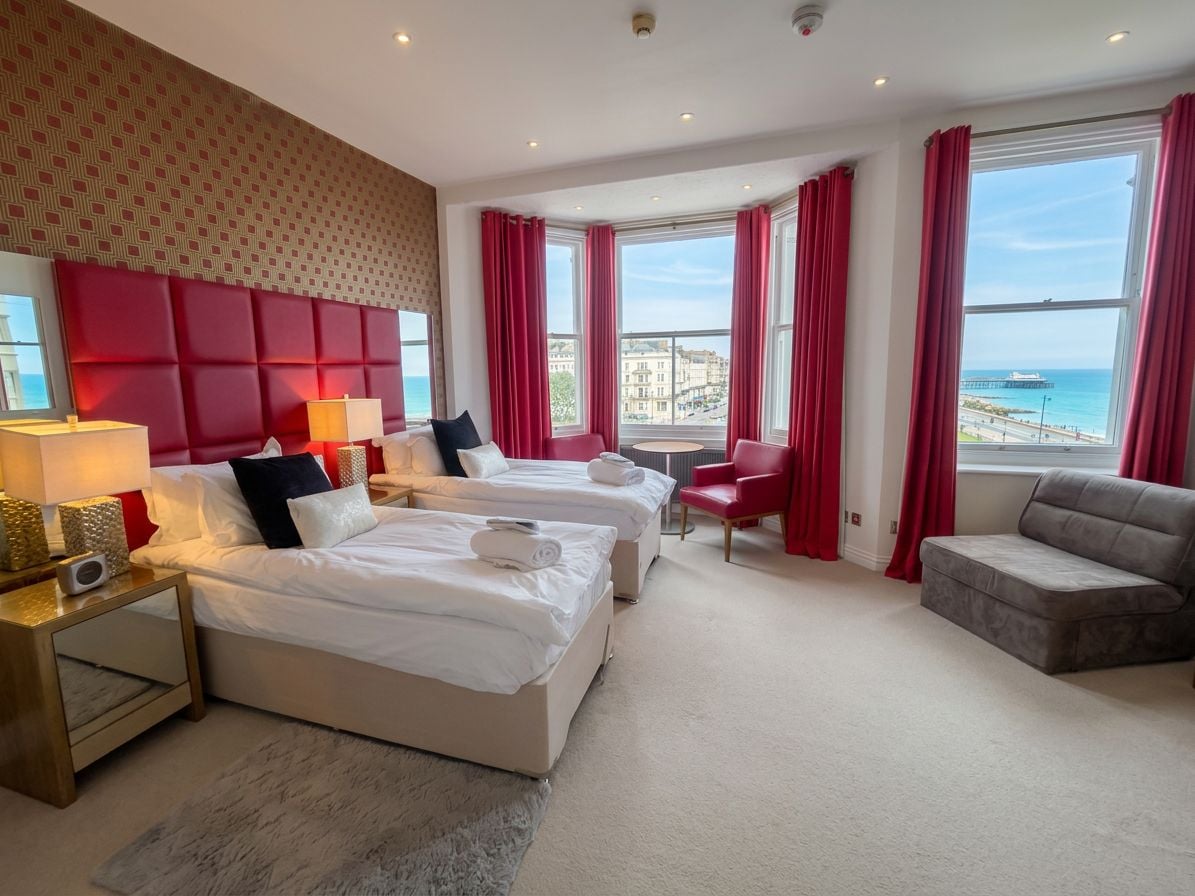 A hotel room with two beds, a red chair, a grey sofa, and a large window with a view of the sea.