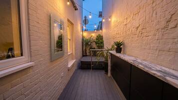 A narrow outdoor corridor with a wooden deck, brick walls, and string lights.