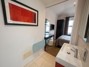 A hotel room with a bed, a bedside table, a TV, a bathroom with a sink and mirror, and a large window with curtains. - Hastings House (St Leonards-On-Sea)