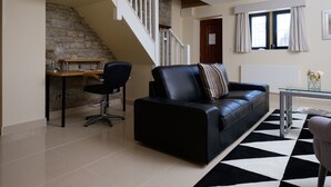 A modern living room with a black leather sofa, a glass coffee table, and a black and white rug. There is a stone wall and a wooden desk with a chair. A staircase is visible in the background. - The Great House Hotel & Restaurant (Bridgend)