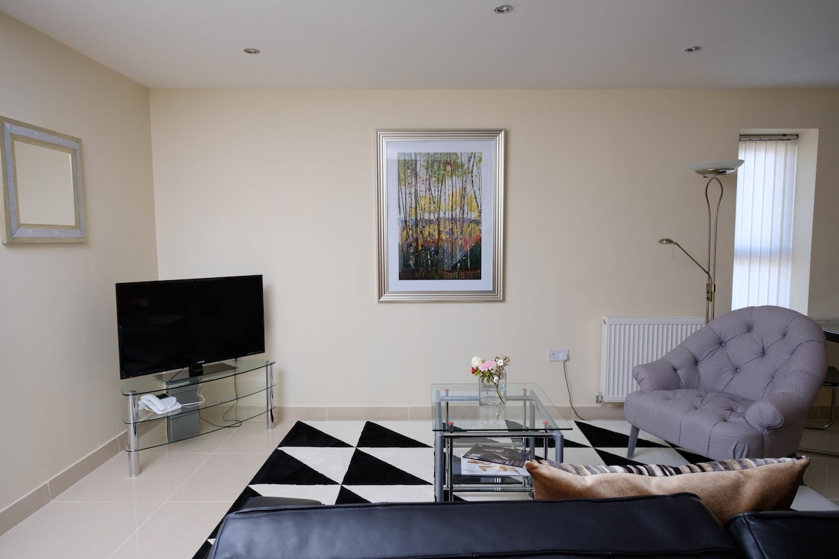 A living room with a flat-screen TV, a glass coffee table with a vase of flowers, a tufted armchair, and a black and white patterned rug.
