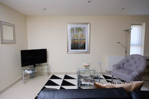 Uma sala de estar com televisão de tela plana, mesa de centro de vidro com um vaso de flores, poltrona estofada e um tapete estampado em preto e branco.