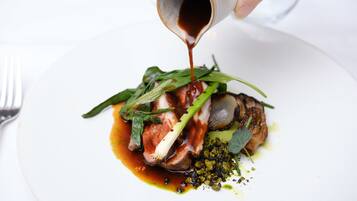 A hand pouring sauce over a dish with meat and greens.