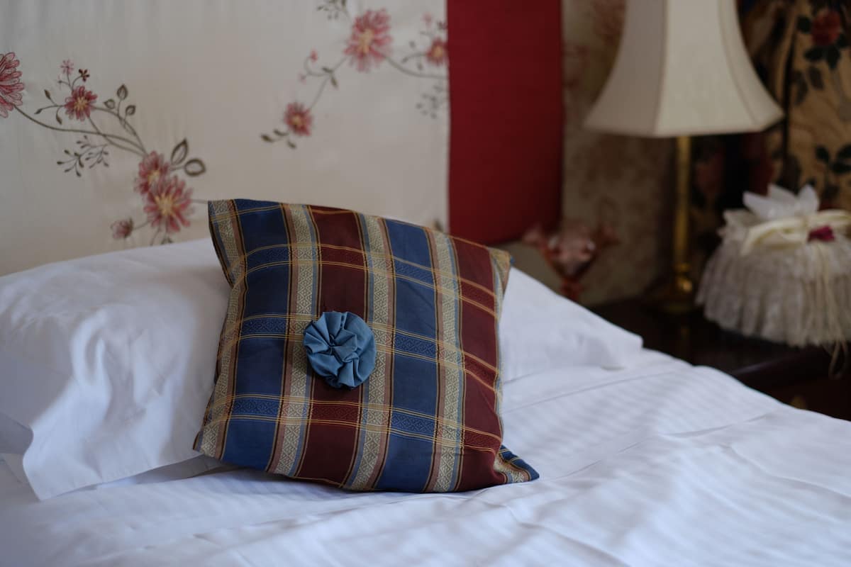 A bed with a plaid pillow and a decorative flower, a lamp, and a floral wallpaper.
