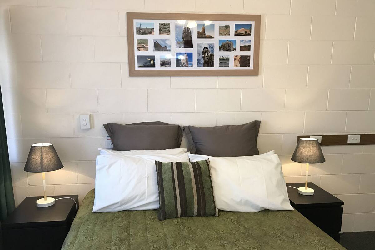 A hotel room with a bed, two nightstands, two lamps, a framed collage on the wall, and a green patterned bedspread.