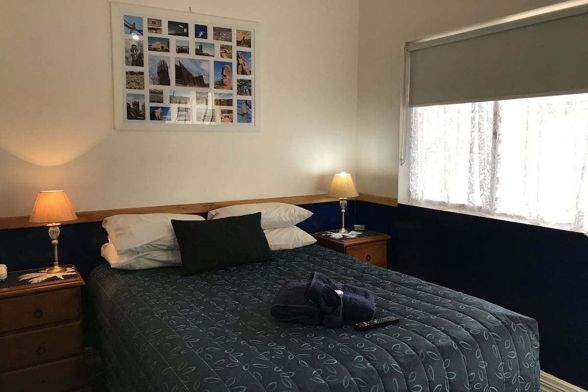 A neatly made bed with a dark blue quilt, two bedside lamps, a wooden nightstand, a dresser, and a wall-mounted photo collage.