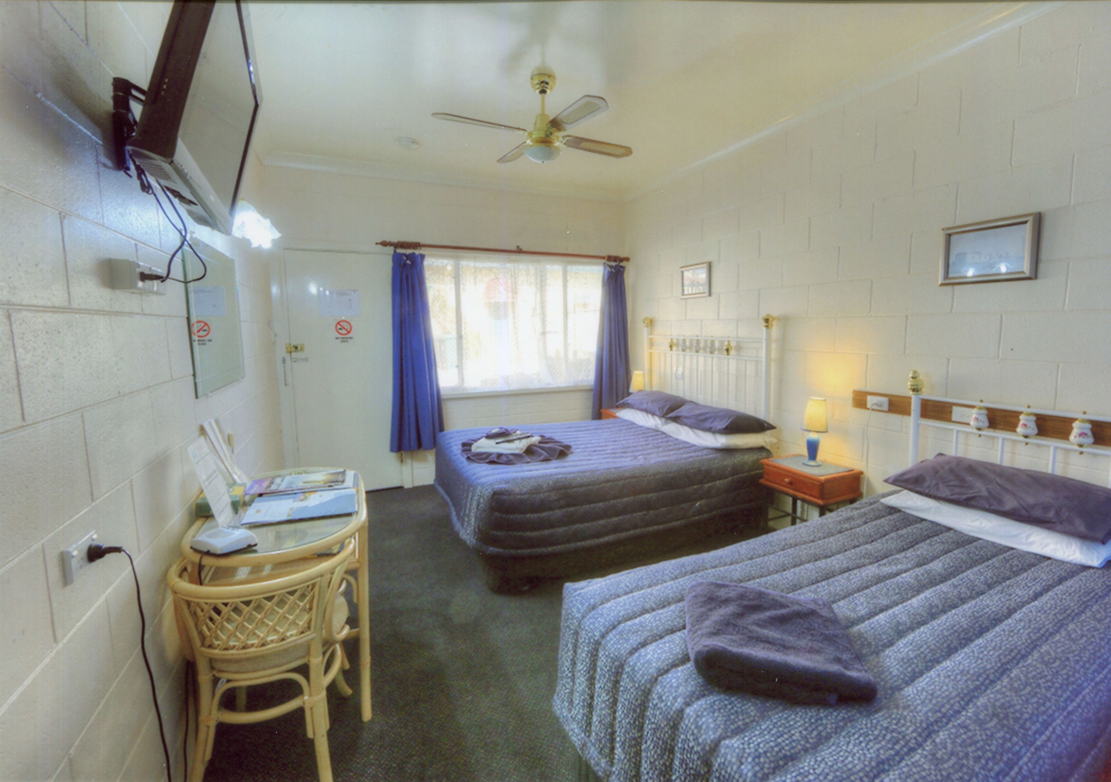 Une chambre d’hôtel avec deux lits, un bureau, une chaise, un ventilateur de plafond et une fenêtre avec des rideaux.