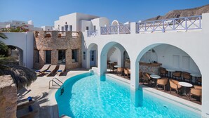 Ein Poolbereich mit Liegestühlen und Tischen, ein Gebäude mit Arkaden und einem strahlend blauen Himmel.