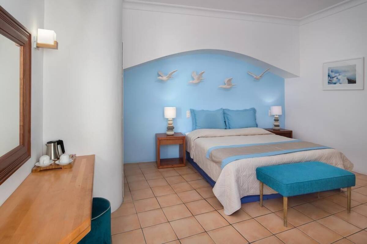 Une chambre à coucher comprenant un lit, des tables de chevet, un banc, un miroir et un mur orné d’une arche bleue et de motifs d’oiseaux.