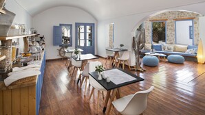 Ein Essbereich mit Holztischen und weißen Stühlen, eine Küche mit Regalen und Gläsern sowie eine gemütliche Sitzecke mit einer Couch und Hocker.