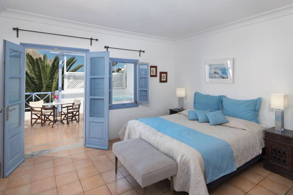 Une chambre à coucher comprenant un lit, des tables de chevet, un banc, une fenêtre et une porte ouverte donnant sur un patio avec une table et des chaises.