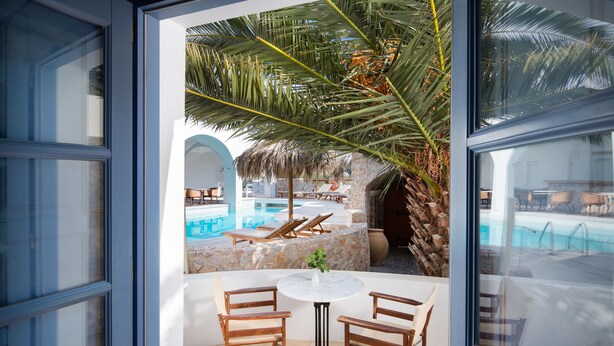 Blick von einem Zimmer mit Glastür auf einen Poolbereich, mit Tisch und Stühlen, einer Palme und Liegestühlen am Pool.