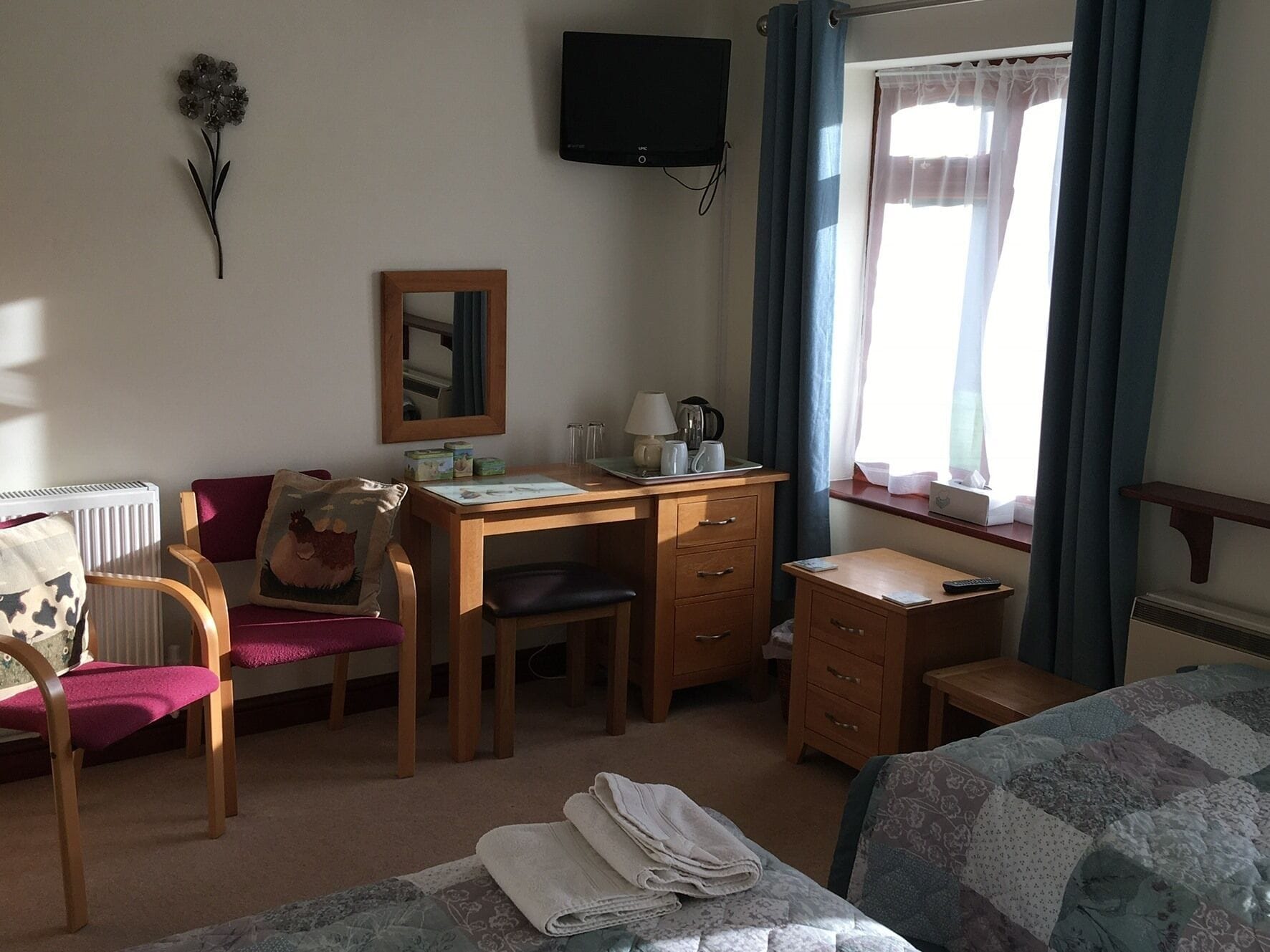 A hotel room with a wooden desk, a chair, a bed with a quilt, a small table, a mirror, a television, and a window with curtains.
