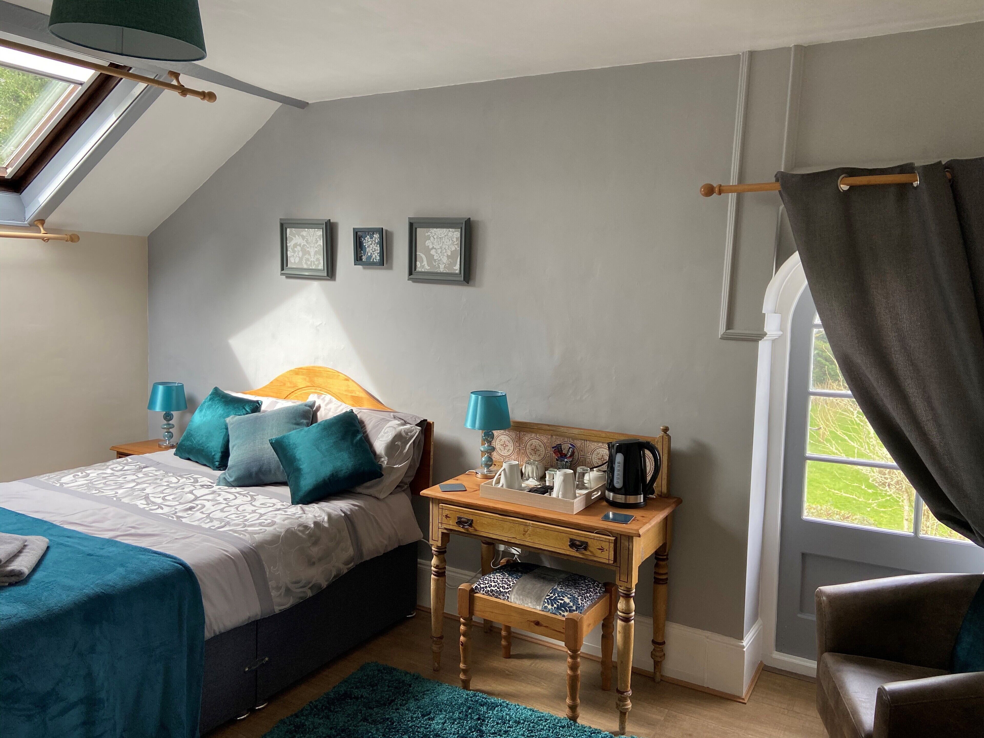 Un dormitorio con cama, mesitas de noche, escritorio, silla, ventana con cortinas y tragaluz.