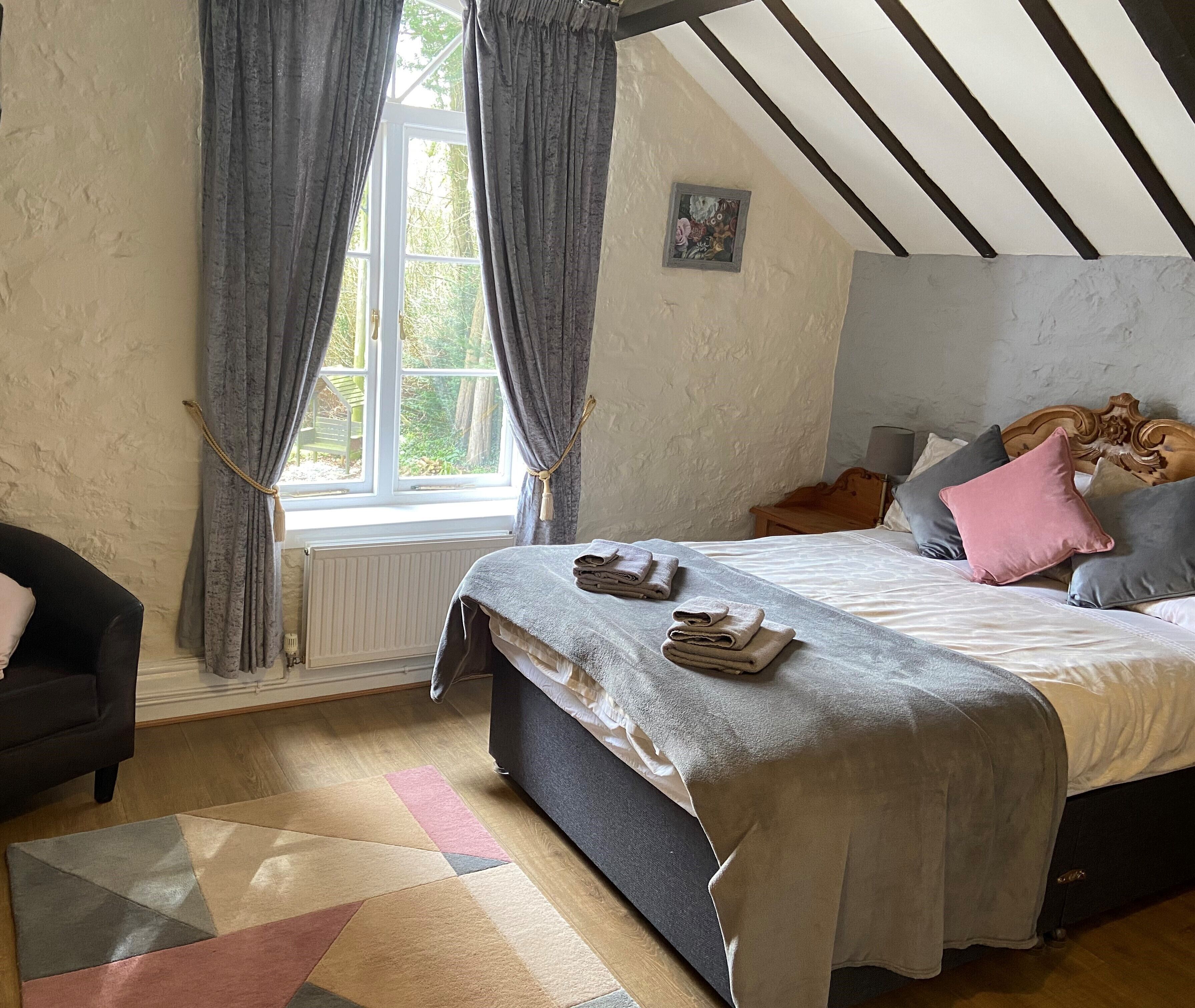 Un dormitorio con cama, una silla, ventana con cortinas, radiador y techo con vigas de madera.
