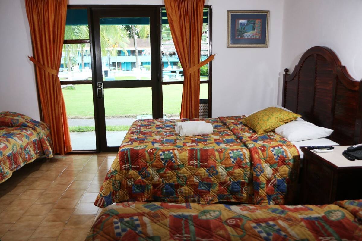A hotel room with two beds, a wooden headboard, a desk with a telephone, and a view of a pool and buildings outside.