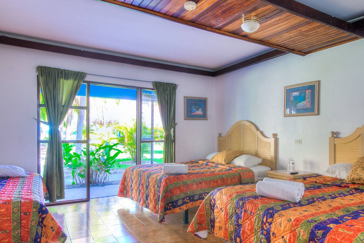 A hotel room with two beds, a large sliding glass door leading to a garden, and a wooden ceiling.