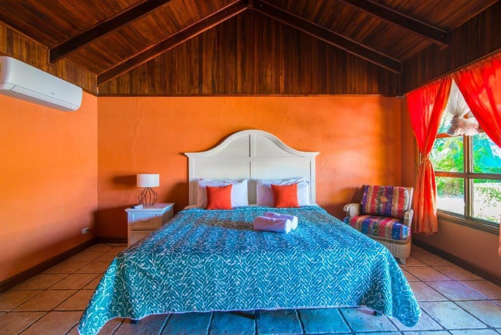 Une pièce avec un plafond en bois, un lit doté d’une tête de lit blanche, des murs orange, une couvre-lit bleue à motifs, une chaise avec un coussin coloré et une fenêtre avec des rideaux rouges.